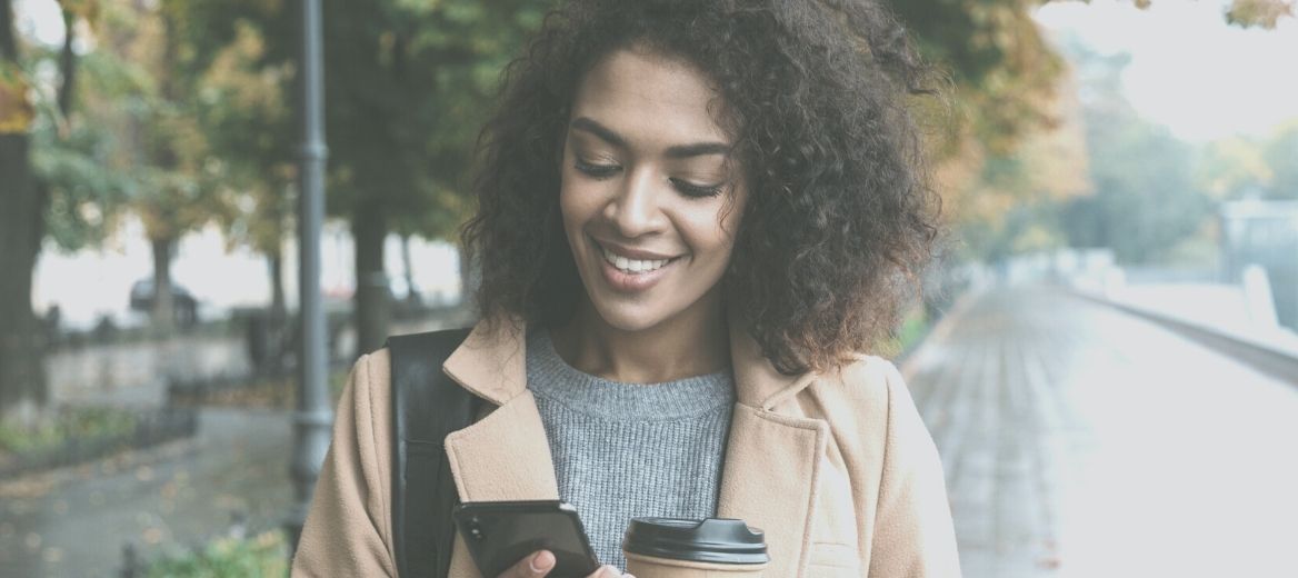 Black woman holding phone smiling wearing brown jacket smiling on a rainy day